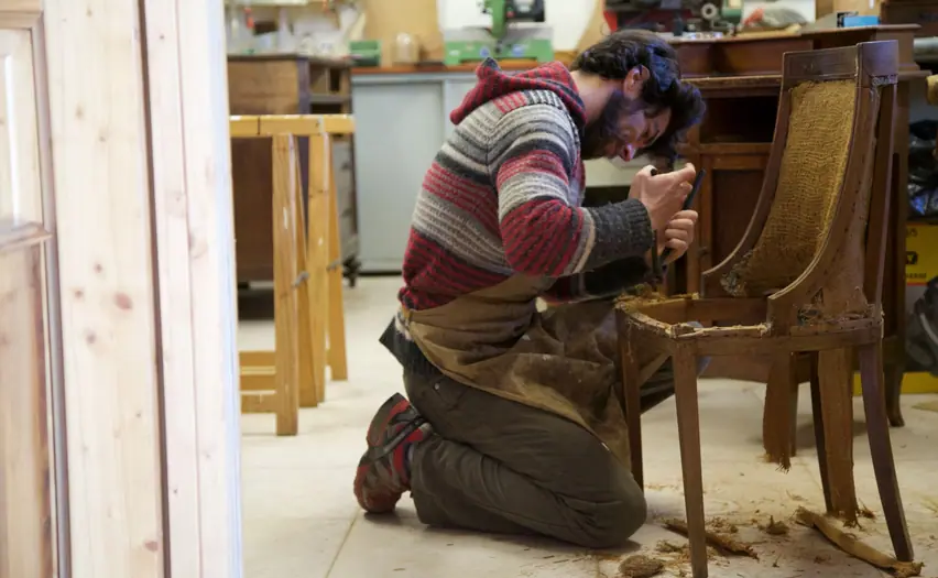 A craftsman kneeling to repair an old wooden chair (stock photo)