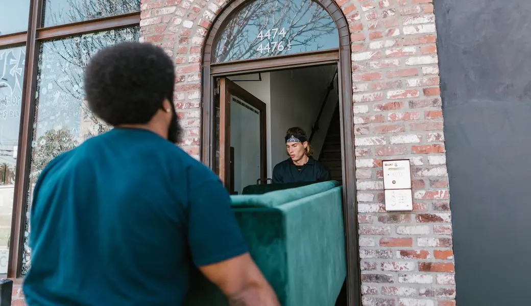 Two movers carrying a green sofa through a brick doorway